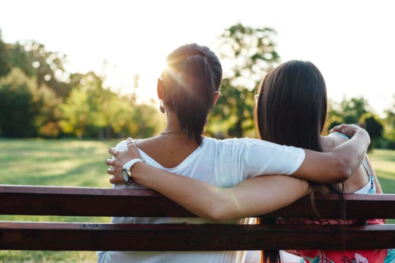 two young woman with their arms around each other's shoulders sitting on a park bench on a sunny day